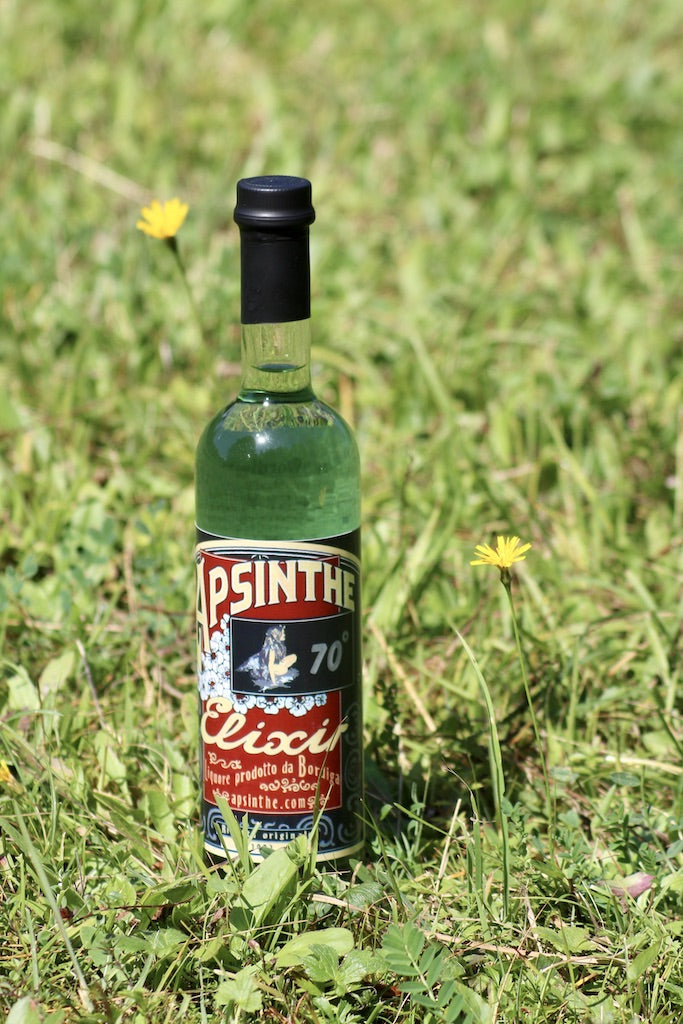 Grüne Absinth-Flasche von Bordiga mit rotem Etikett steht auf einer Wiese neben kleinen gelben Blumen.