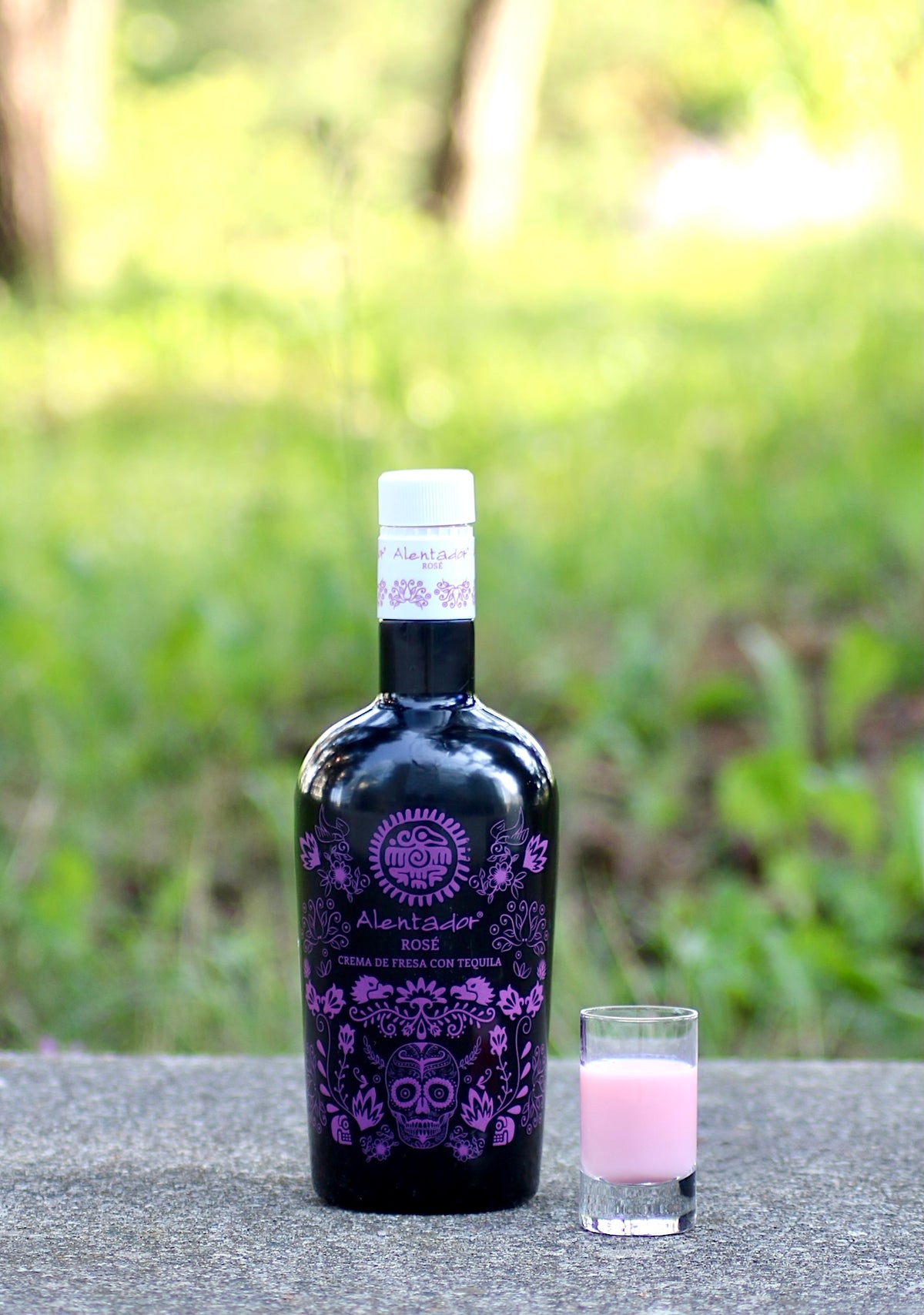 Schwarze Flasche Alentador Rosé Crema Likör mit lila Design, daneben ein gefülltes Shotglas auf grauer Oberfläche