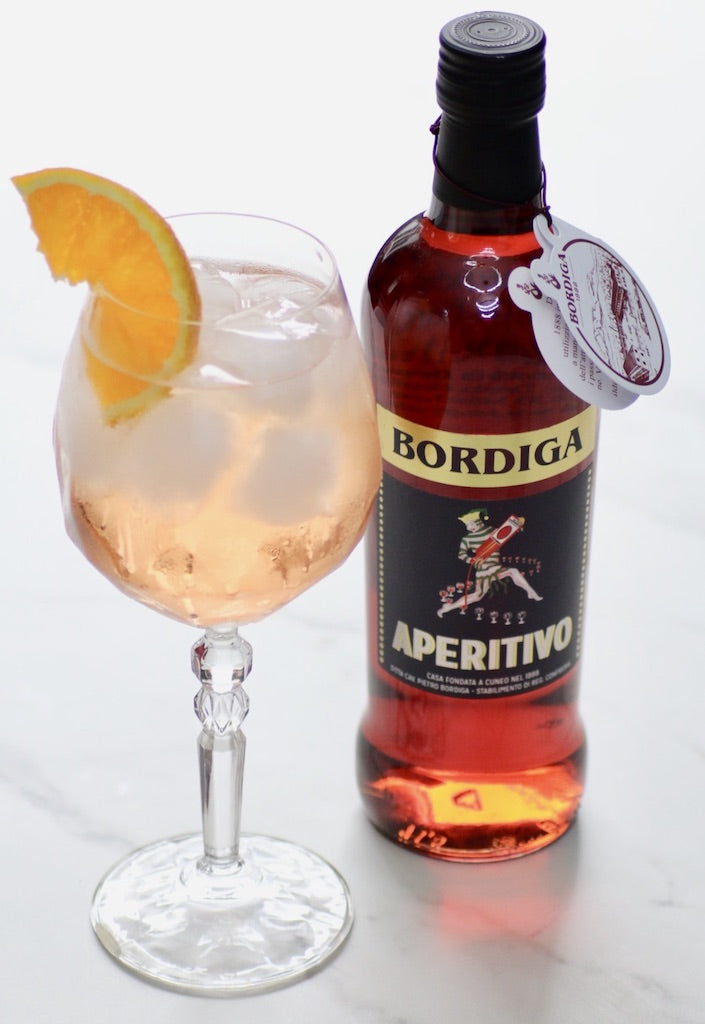 Aperitivo Spritz im geschliffenen Weinglas mit Eis und Orangenscheibe neben einer Flasche Bordiga Aperitivo auf hellem Untergrund.