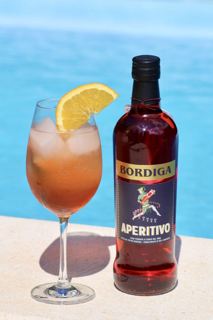 Weinglas mit Aperitivo-Cocktail und Orangenscheibe neben einer Flasche Bordiga Aperitivo am Beckenrand mit Pool im Hintergrund.