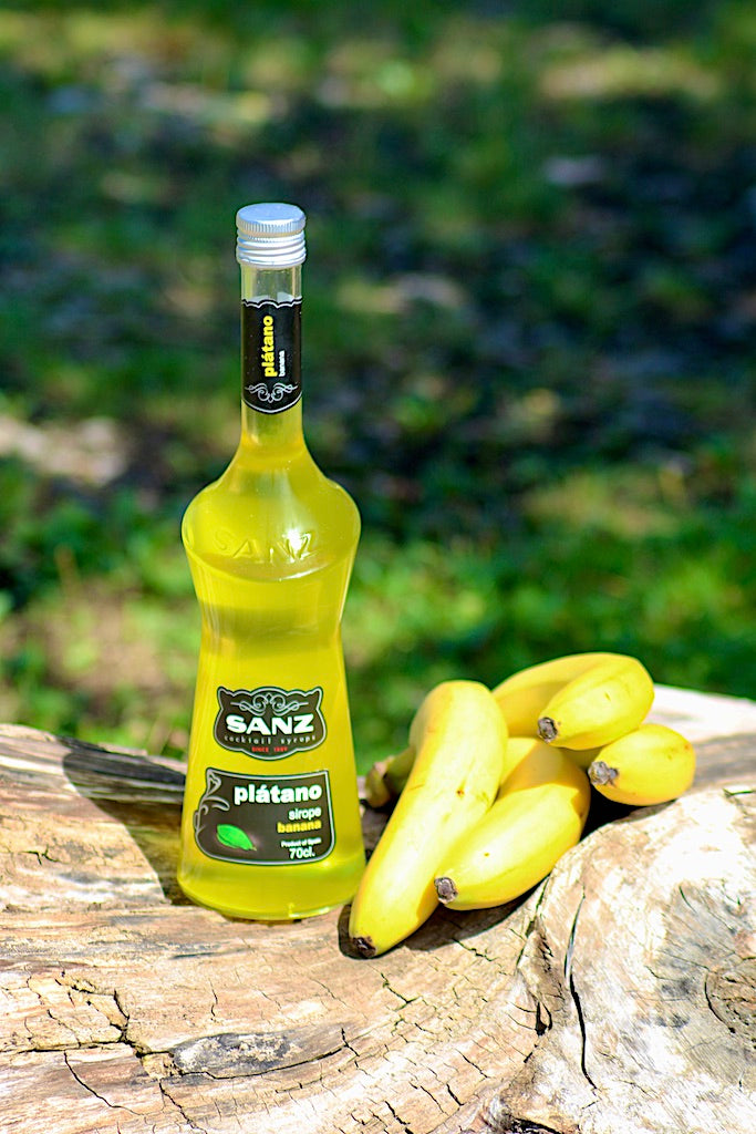 Flasche Sanz Plátano (Banane) Sirup, gelber Sirup in 700 ml Flasche, neben Bündel frischer Bananen auf einem Baumstamm.