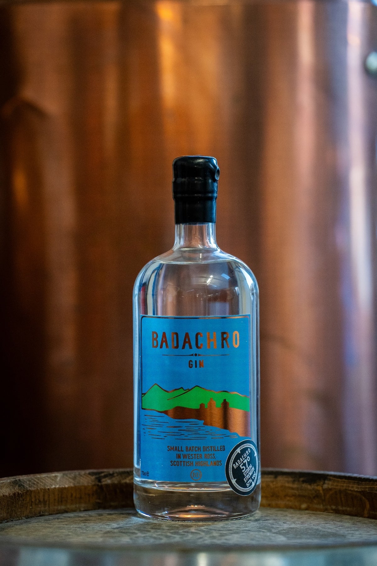 Flasche Badachro Storm Strength Gin auf einem Holzfass vor einem Kupfer-Destillationsapparat in der Destillerie.