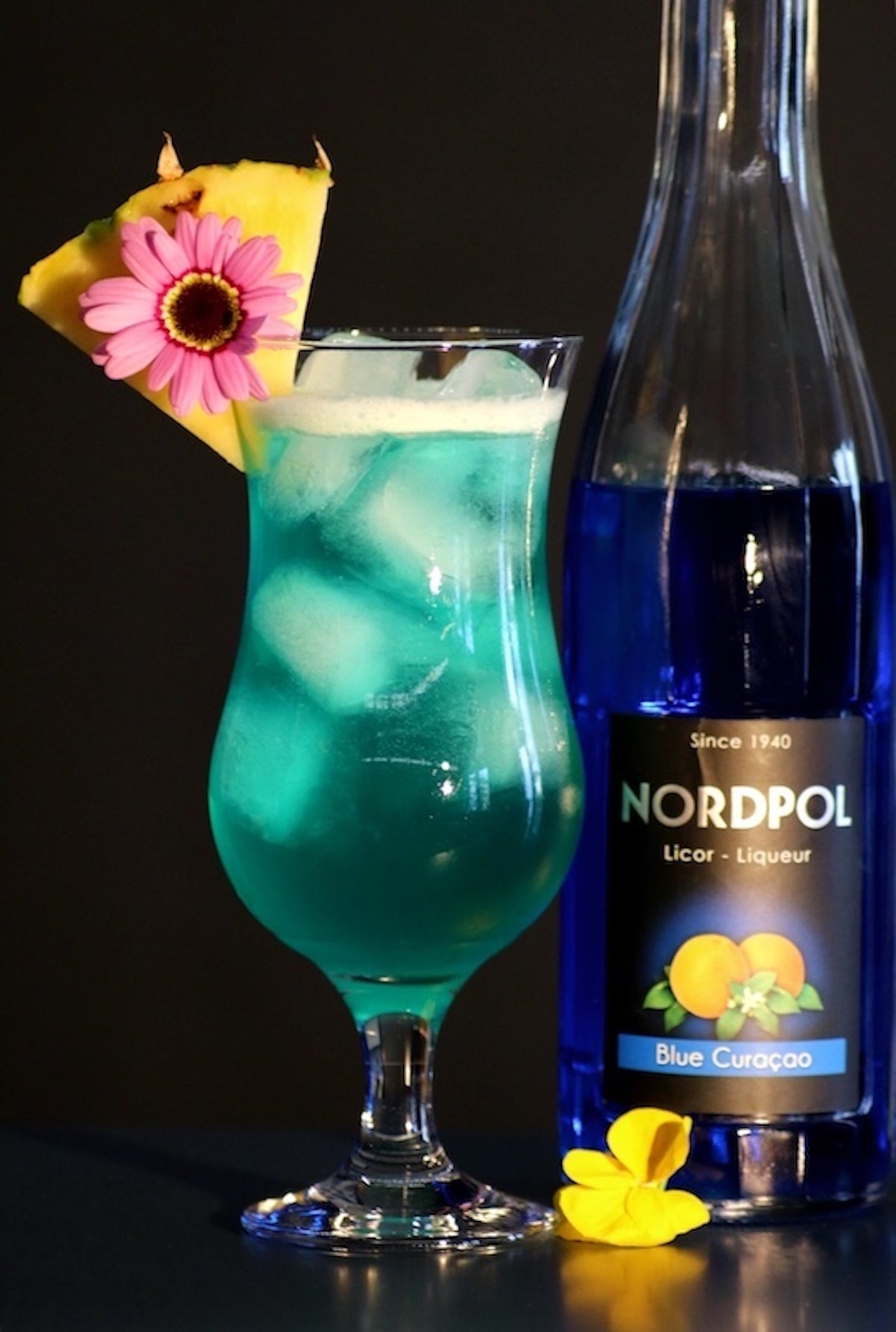 Blue Hawaii Cocktail mit Eiswürfeln, Ananasscheibe und Blüte, daneben Flasche Blue Curacao von Nordpol.