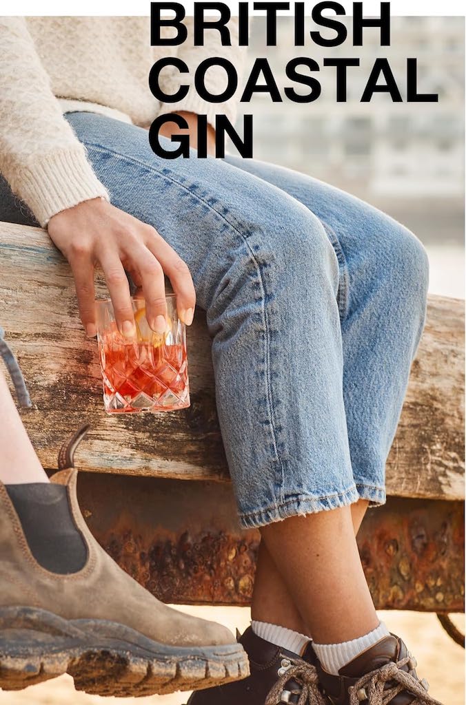Person sitzt auf Holzbank am Wasser mit rotem Cocktail im Glas, Text „British Coastal Gin“ im Bild