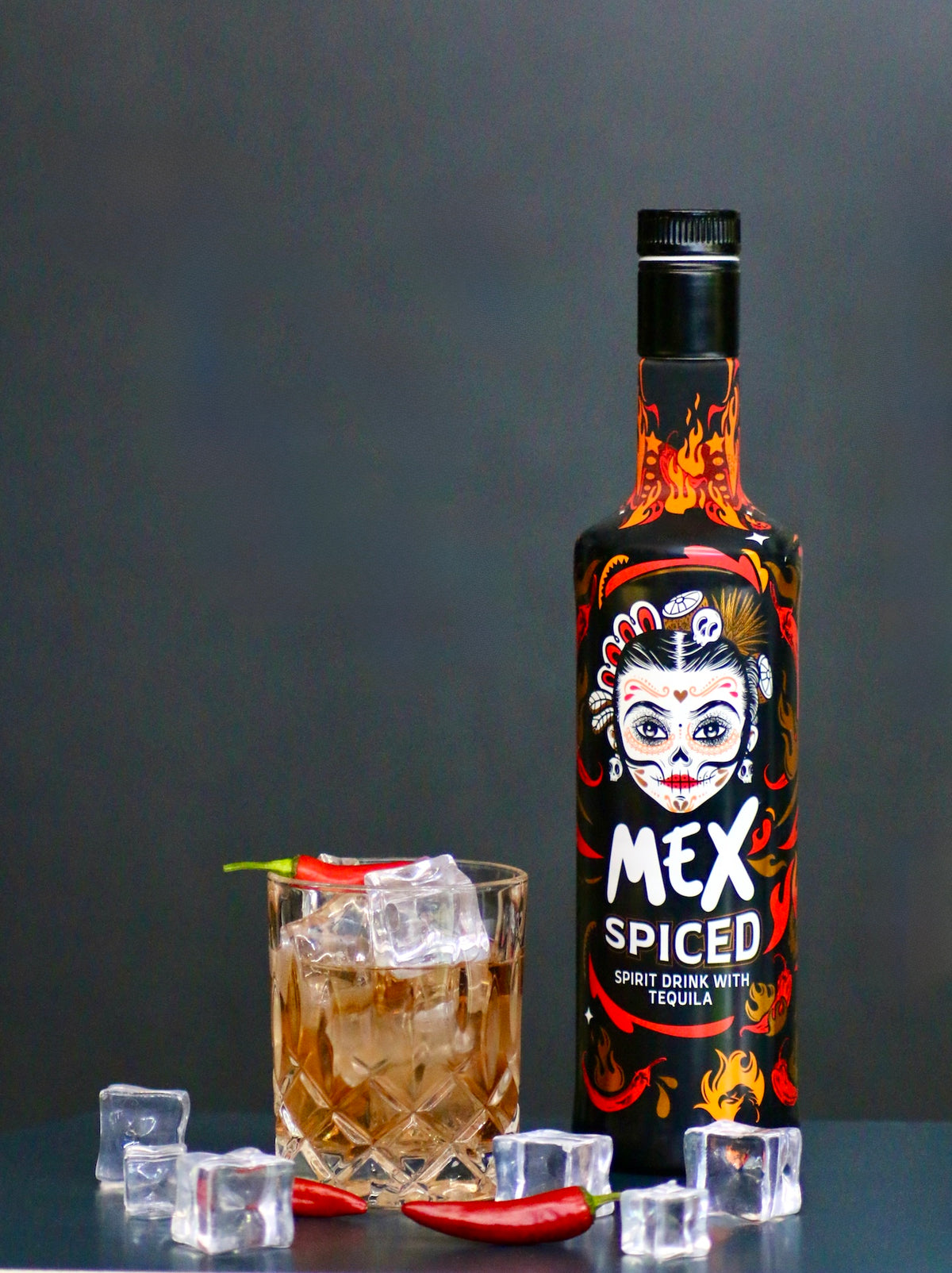 Burning Sunset Cocktail mit Eiswürfeln und Chili, serviert im Tumbler, neben der Flasche Mex Spiced mit feurigem Flammen-Design.