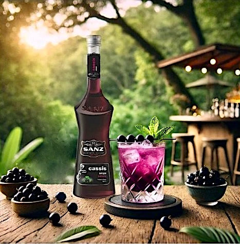 Flasche Cassis-Sirup von Sanz neben einem violetten Cocktail mit Eis, Minze und schwarzen Johannisbeeren.