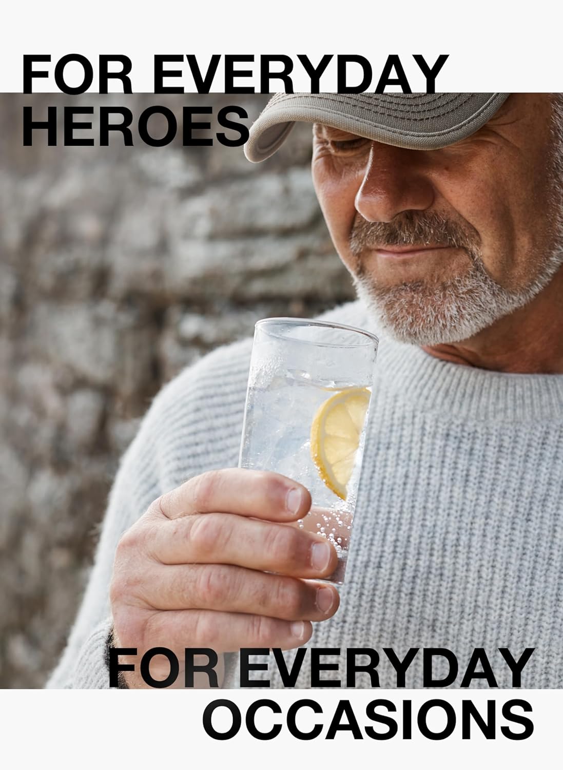 Mann hält Gin Tonic mit Zitronenscheibe im Glas, Nahaufnahme vor Steinwand, Text „For everyday heroes“ im Bild