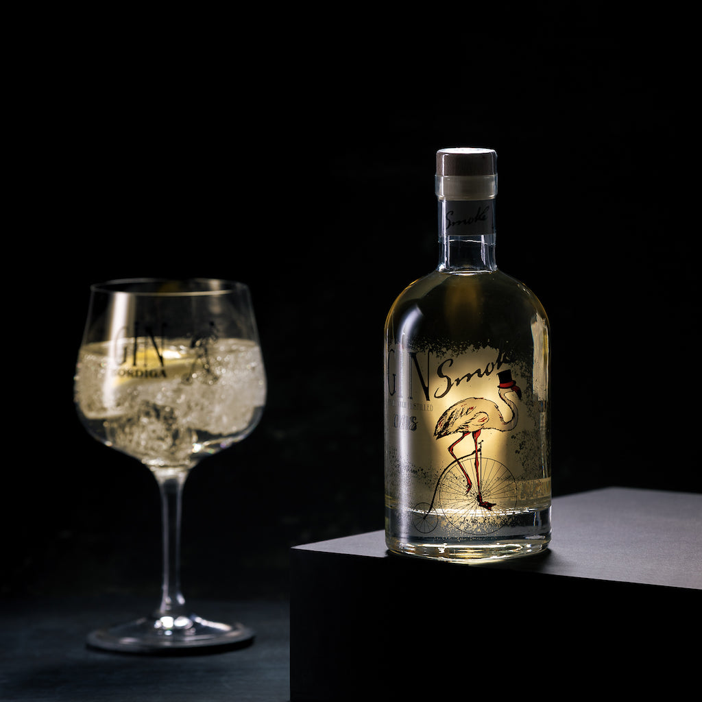 Flasche Ciais Gin Smoke von Bordiga auf Podest, daneben ein Ballonglas mit Gin Tonic und Eiswürfeln vor schwarzem Hintergrund.