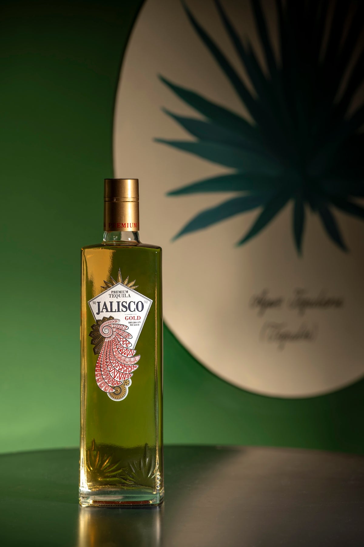Tequila-Jalisco-Gold-Flasche auf dunkler Fläche vor grünem Hintergrund mit großem Agavenmotiv.