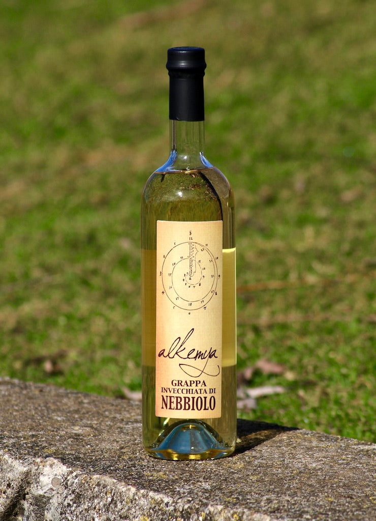 "Flasche Grappa Invecchiata di Nebbiolo mit goldfarbenem Etikett, stehend auf einer Mauer vor einer grünen Wiese.