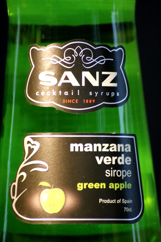 Nahaufnahme des Frontetiketts von Sanz Grüner Apfel Sirup mit weißem Logo und grünem Apfelsymbol auf schwarzem Hintergrund.