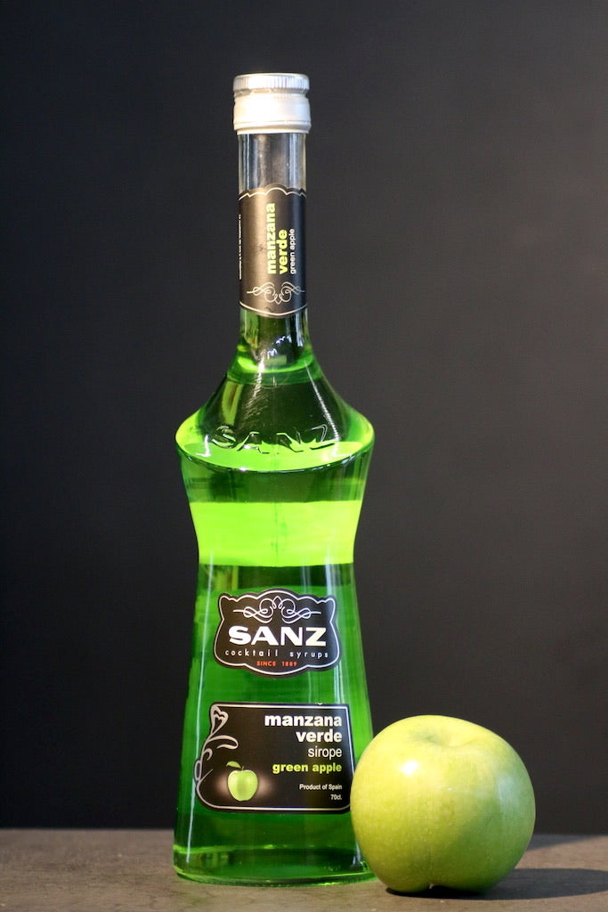 Flasche Grüner Apfel Sirup von Sanz mit leuchtend grünem Inhalt, schwarzem Etikett und einem grünen Apfel davor.