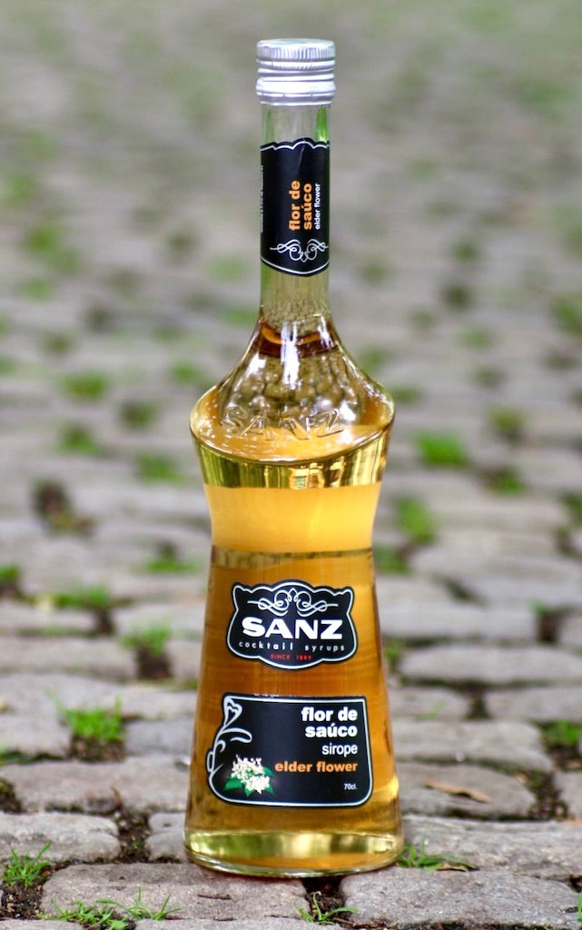 Holunderblütensirup Sanz 0,7L auf Kopfsteinpflaster mit natürlichem Licht und grünem Moos zwischen den Steinen.