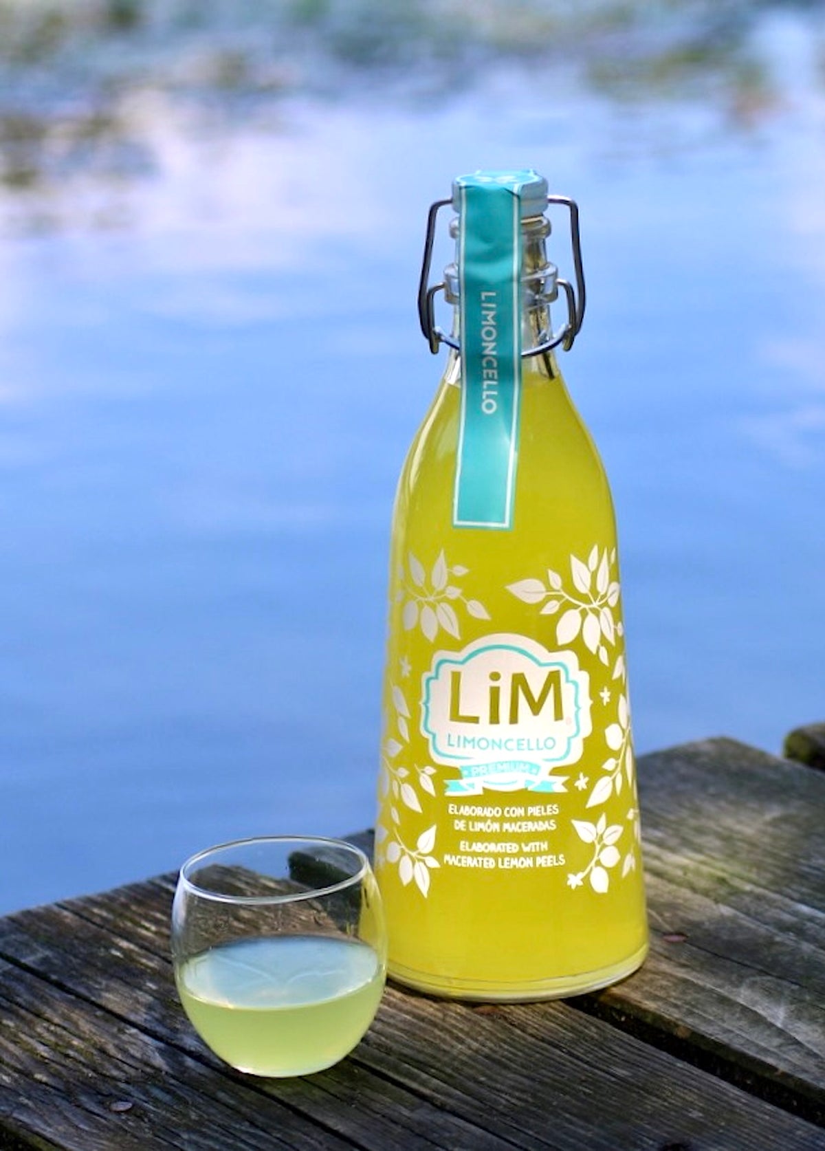 Lim Limoncello Flasche mit kleinem Glas auf Holzsteg vor blauem Wasser im Hintergrund