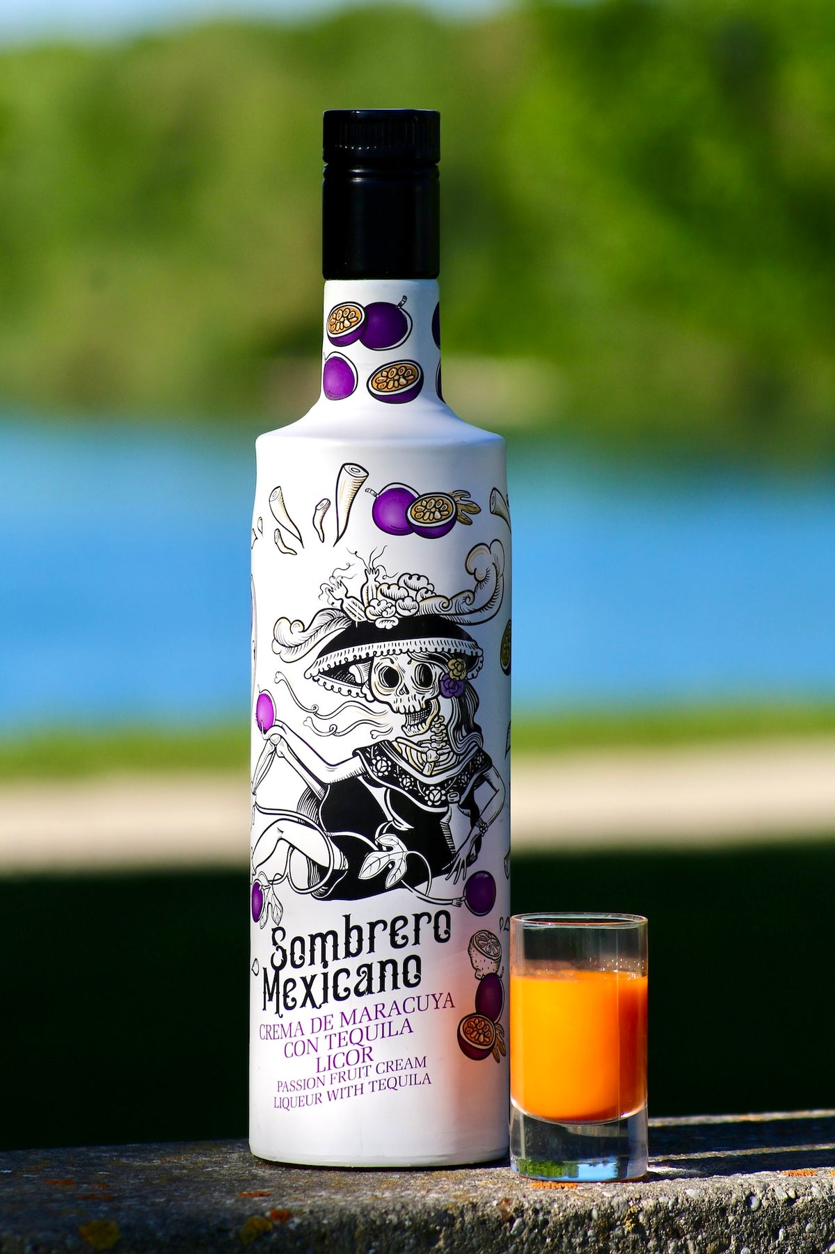 Flasche Sombrero Mexicano Maracuja-Tequila-Likör mit orangem Shotglas, vor See und grünem Hintergrund auf Steinfläche.