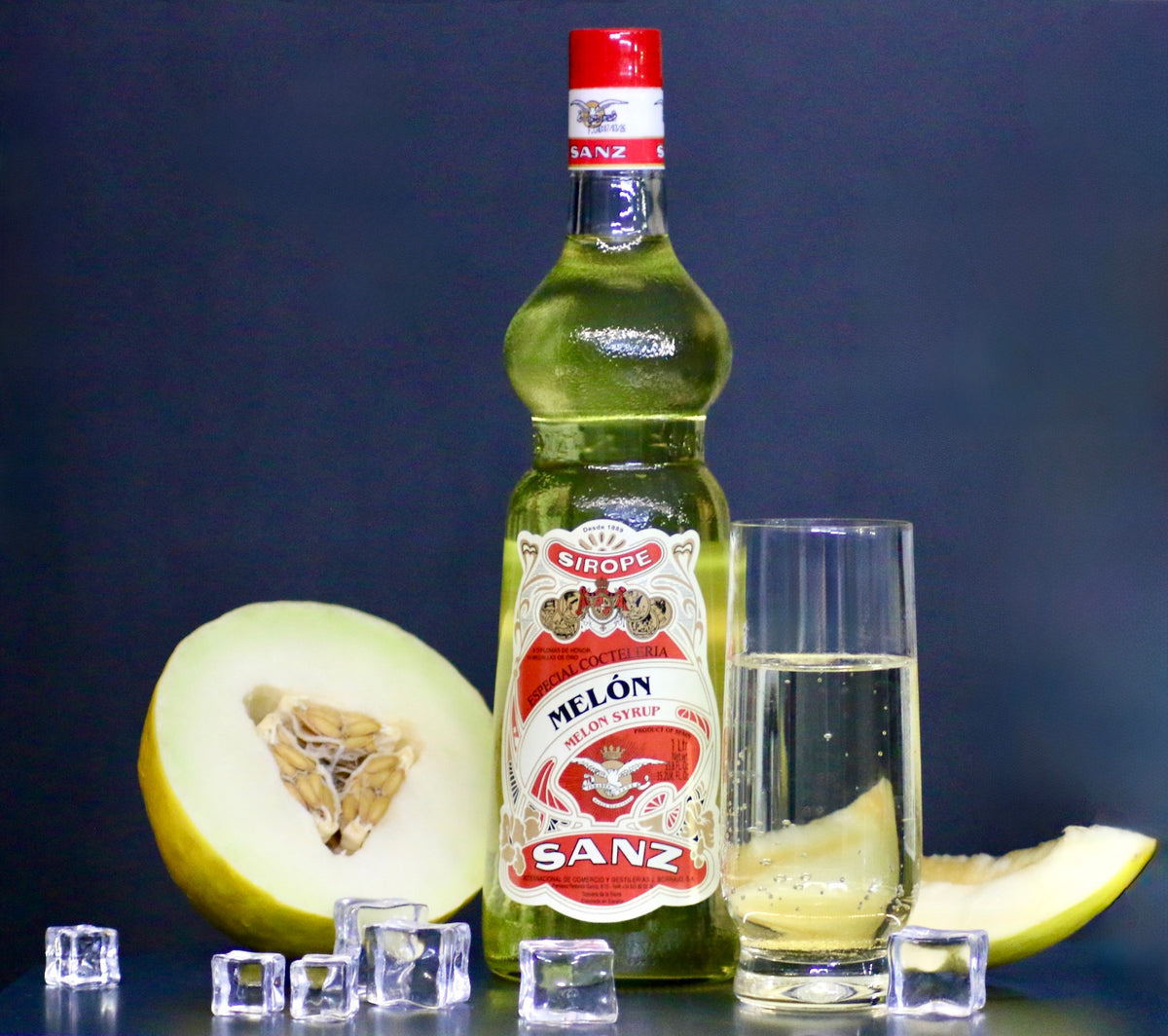 Melonensirup Sanz 1L, gelber Sirup in Flasche mit rotem Etikett, Glas mit Sirup, Melonenscheiben und Eiswürfeln.