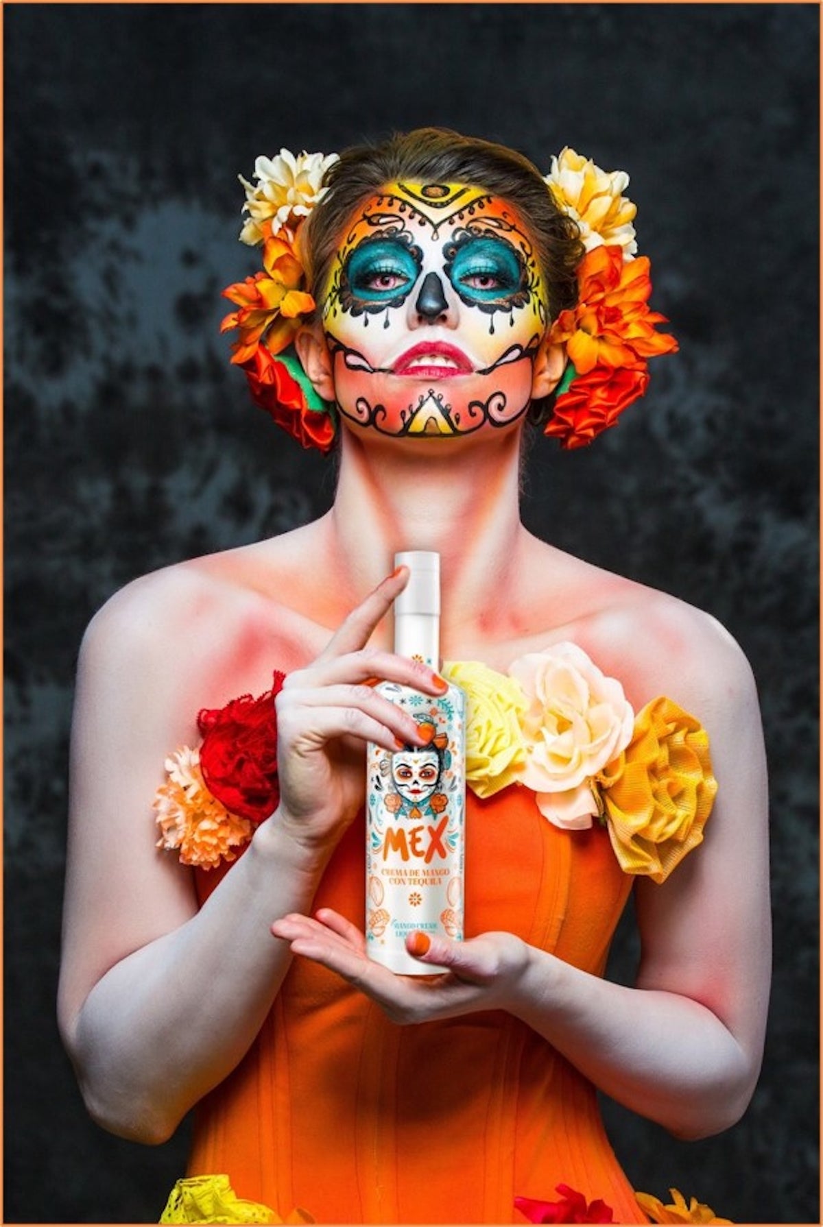 Frau im Catrina-Stil mit farbenfrohem Make-up und Blumen hält eine Flasche Mex Mango-Tequila Likör mit Sahne vor dunklem Hintergrund.