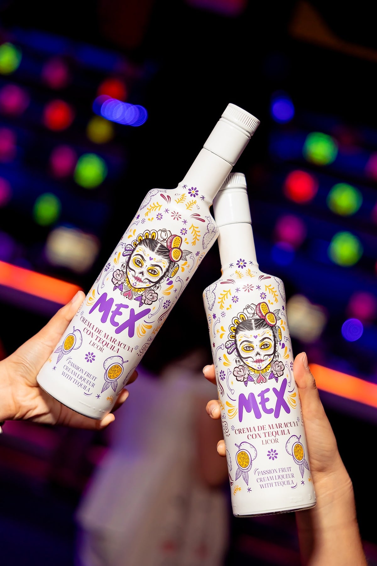 Zwei Flaschen Mex Maracuja-Tequila Sahnelikör werden vor buntem Partyhintergrund in die Höhe gehalten.