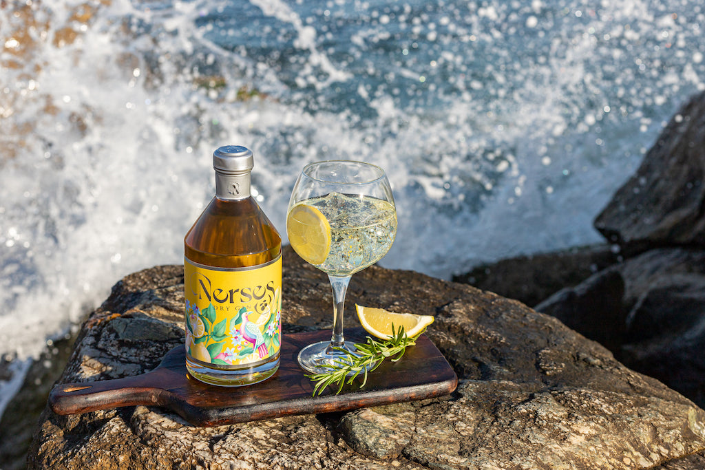 Nerses Dry Gin Flasche neben Gin-Tonic-Glas mit Zitronenscheibe und Rosmarin auf Felsen am Meer mit spritzenden Wellen
