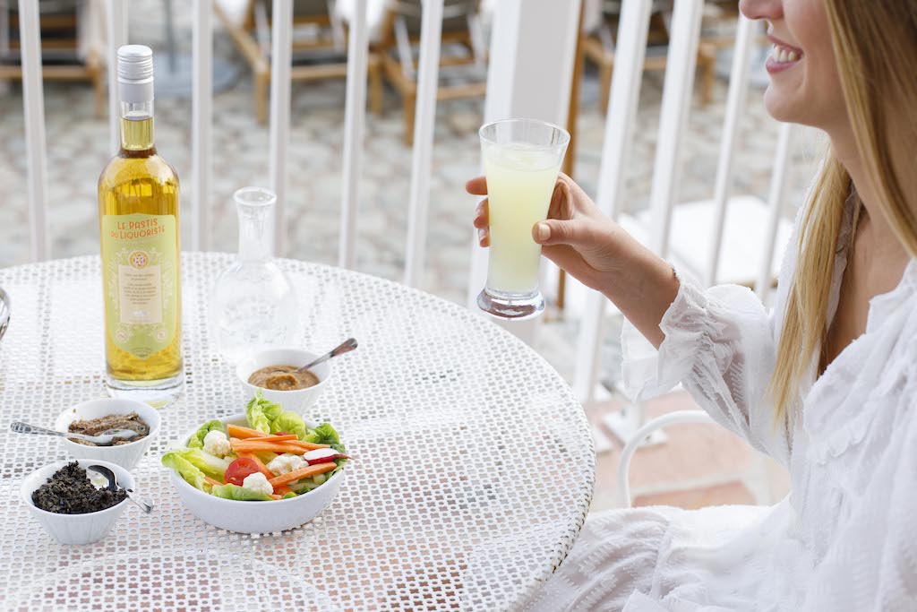 Eine Frau hält ein Glas Pastis neben einer Flasche Le Pastis du Liquoriste, begleitet von Salat und Dips auf einem weißen Tisch.