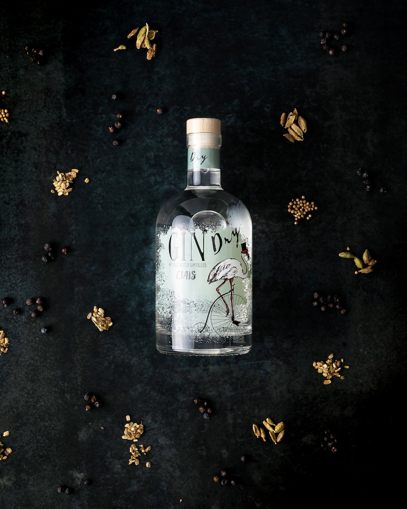 Flasche Bordiga Gin Ciais mit Flamingo-Design auf dunklem Untergrund, umgeben von Wacholderbeeren, Kardamom und weiteren Botanicals.