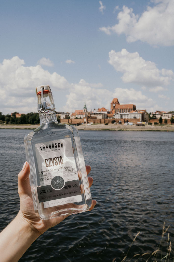 Hand hält Flasche Toruńska Czysta Wodka 40% vol. vor dem Flussufer und der historischen Altstadt von Toruń, Polen.