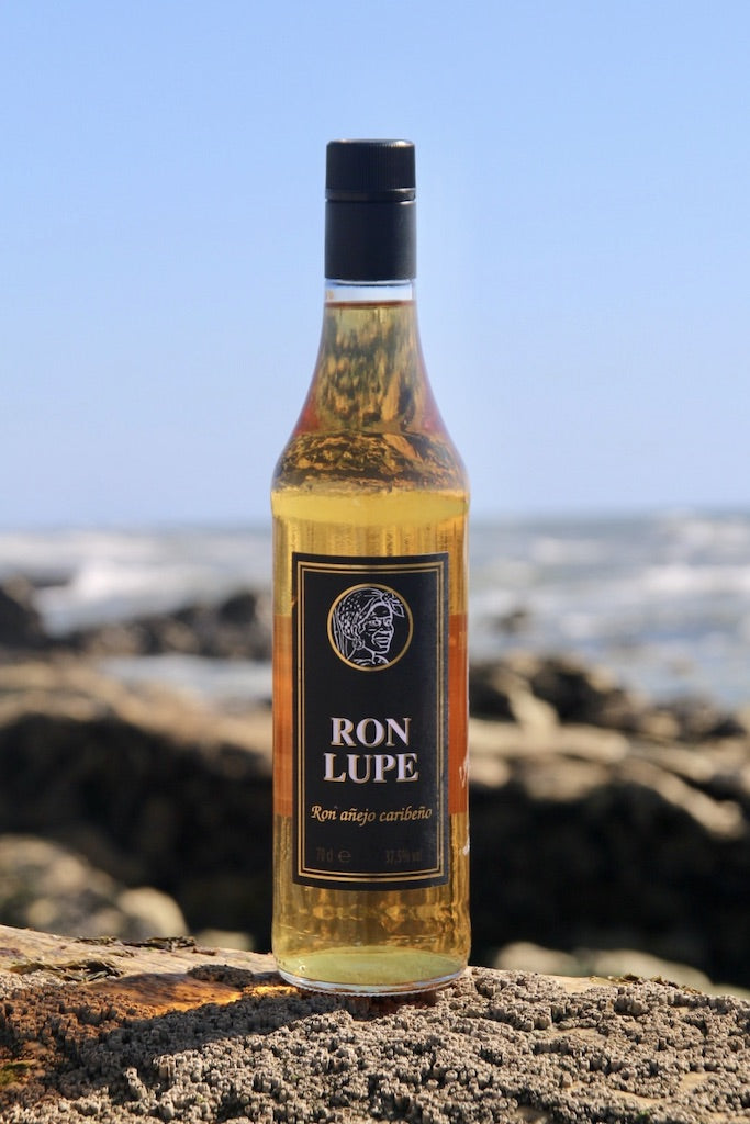 Rum Añejo Lupe, goldbraune Flasche mit schwarzem Etikett, am Meer auf Felsen platziert mit blauem Himmel im Hintergrund.
