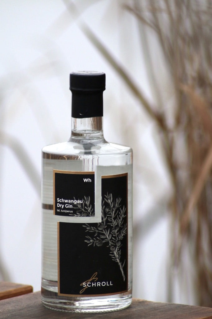 Schwangau Dry Gin Flasche mit schwarzem Etikett steht auf einer Holzfläche, im Hintergrund natürliche Gräser und ein heller Himmel.