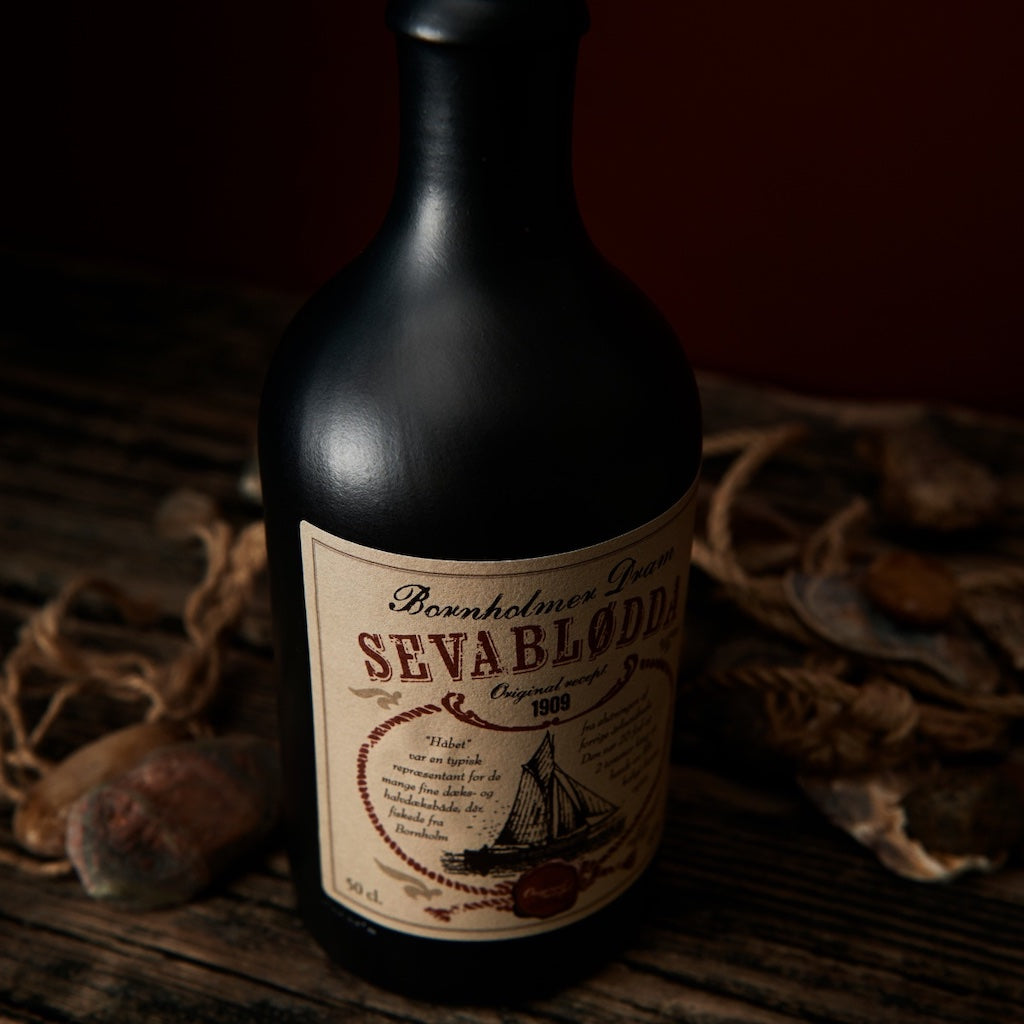 Bornholmer Dram Sevablødda 50cl, schwarze Flasche mit Vintage-Label, auf rustikalem Holzuntergrund mit Seilen und Steinen arrangiert.