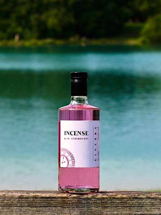 Flasche Incense Gin mit Erdbeergeschmack, rosa Flüssigkeit, auf einem Holzgeländer vor einem See mit Bäumen im Hintergrund.