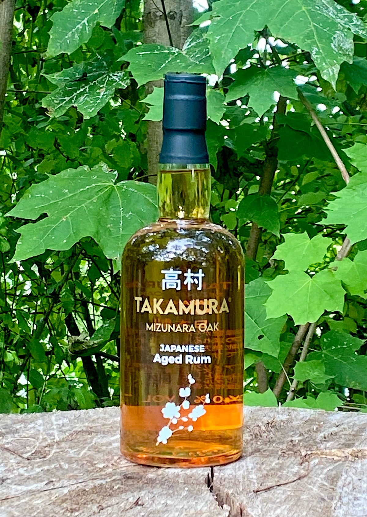 Takamura Mizunara Oak Japanese Aged Rum auf einem Baumstamm, Flasche mit japanischer Schrift und Blütenmotiv.