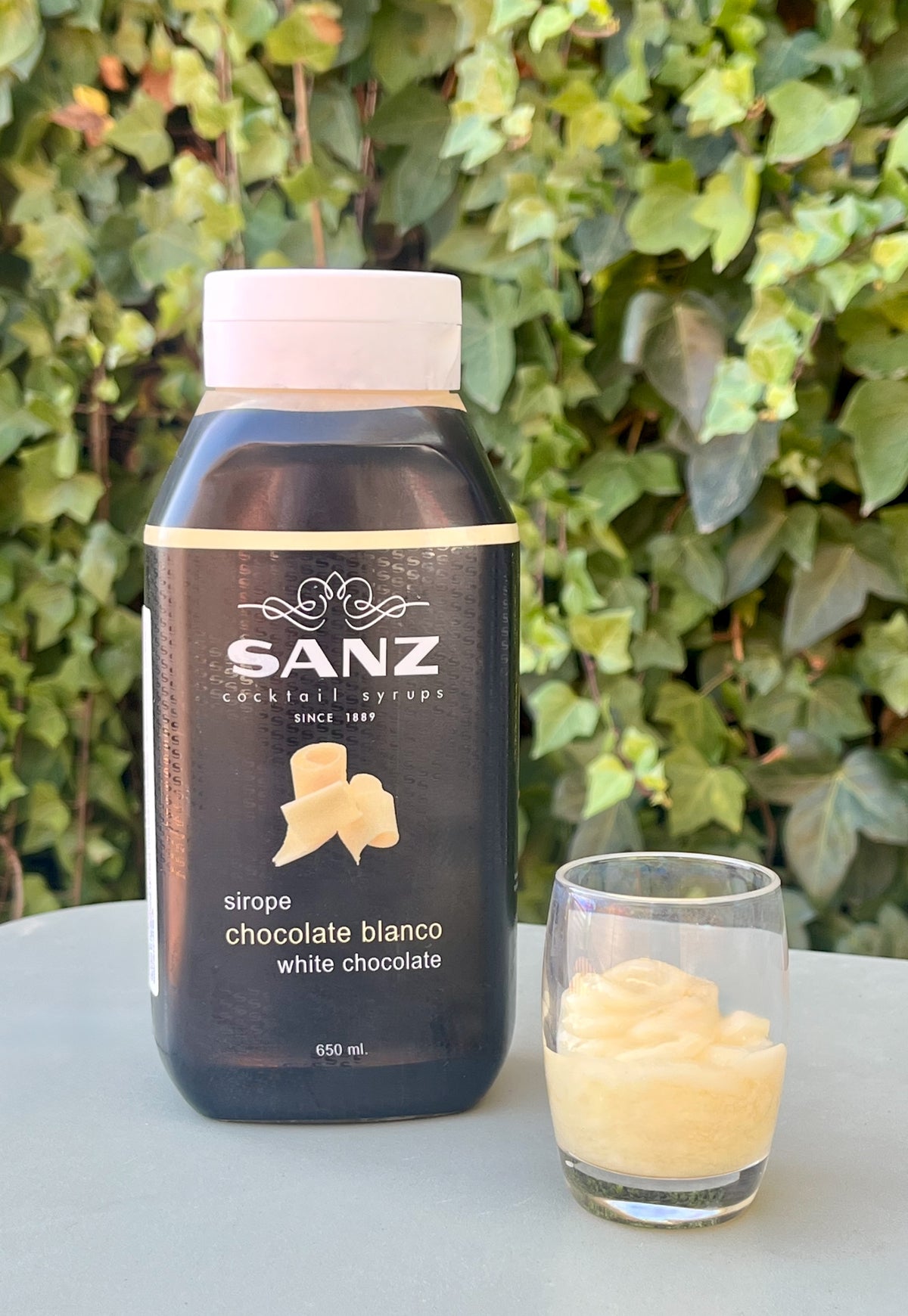 Sanz Sirup Weiße Schokolade mit Flasche und Glas mit hellem Sirup auf einem Tisch vor Efeu.