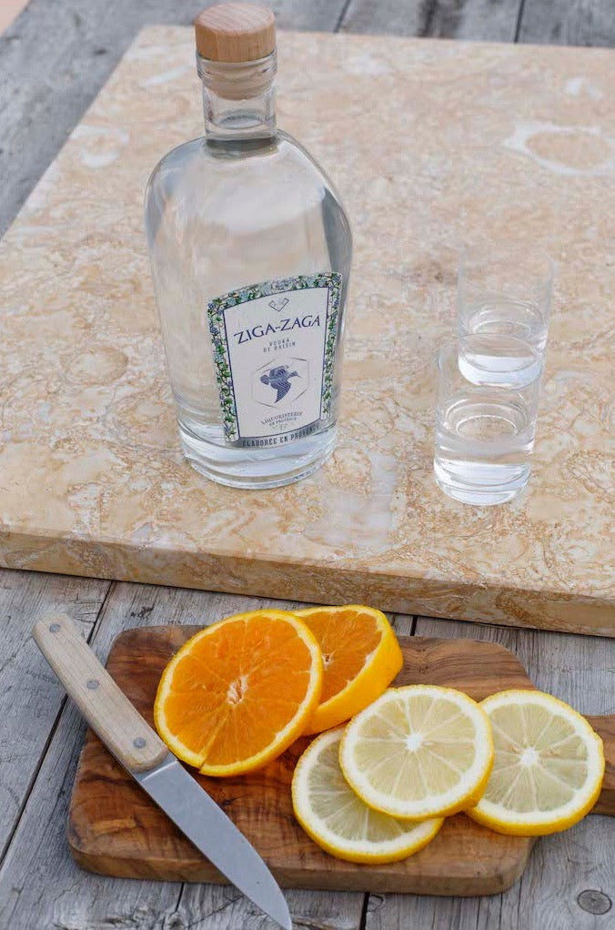 Flasche Ziga Zaga Vodka mit zwei Gläsern auf Steinplatte, davor Orangen- und Zitronenscheiben mit Messer auf Holzbrett.
