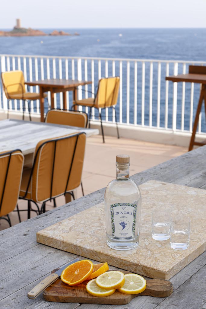 Flasche Ziga Zaga Vodka mit zwei Gläsern und Orangenscheiben auf Terrasse mit Meerblick und Tischen im Hintergrund.