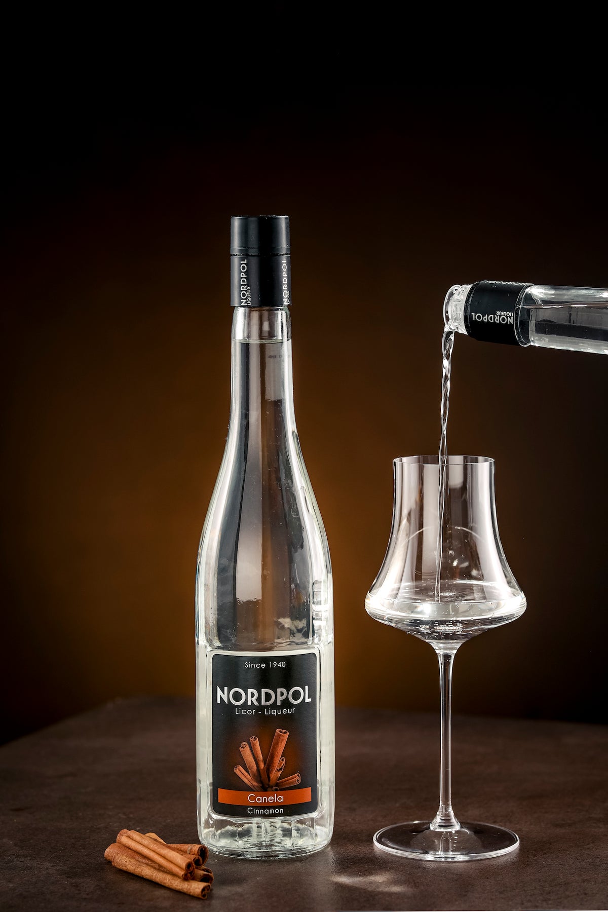 Flasche Nordpol Zimtlikör (Canela) neben Glas, in das Zimtlikör eingeschenkt wird, mit Zimtstangen im Vordergrund auf dunklem Hintergrund.