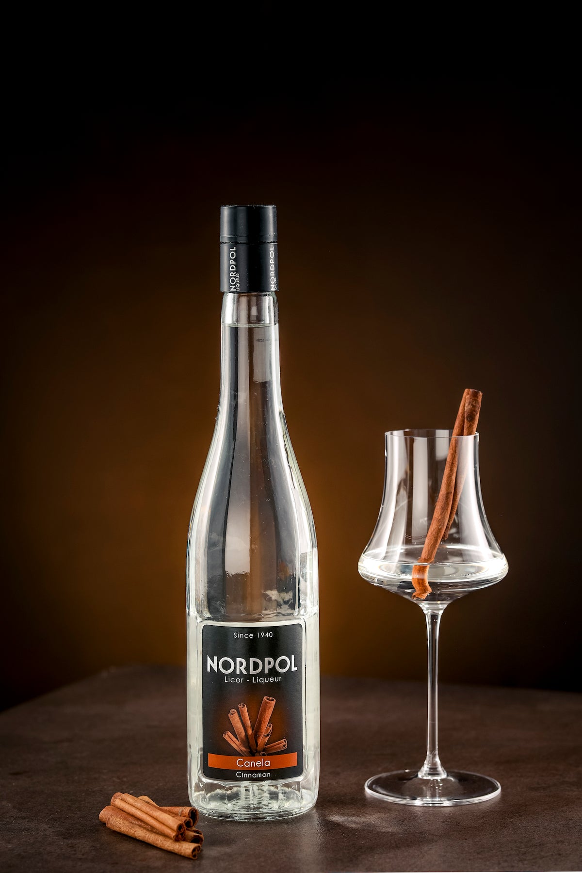 Flasche Nordpol Zimtlikör (Canela) neben Glas mit Zimtstange als Garnitur, vor dunkelbraunem Hintergrund, zusätzliche Zimtstangen vorne.