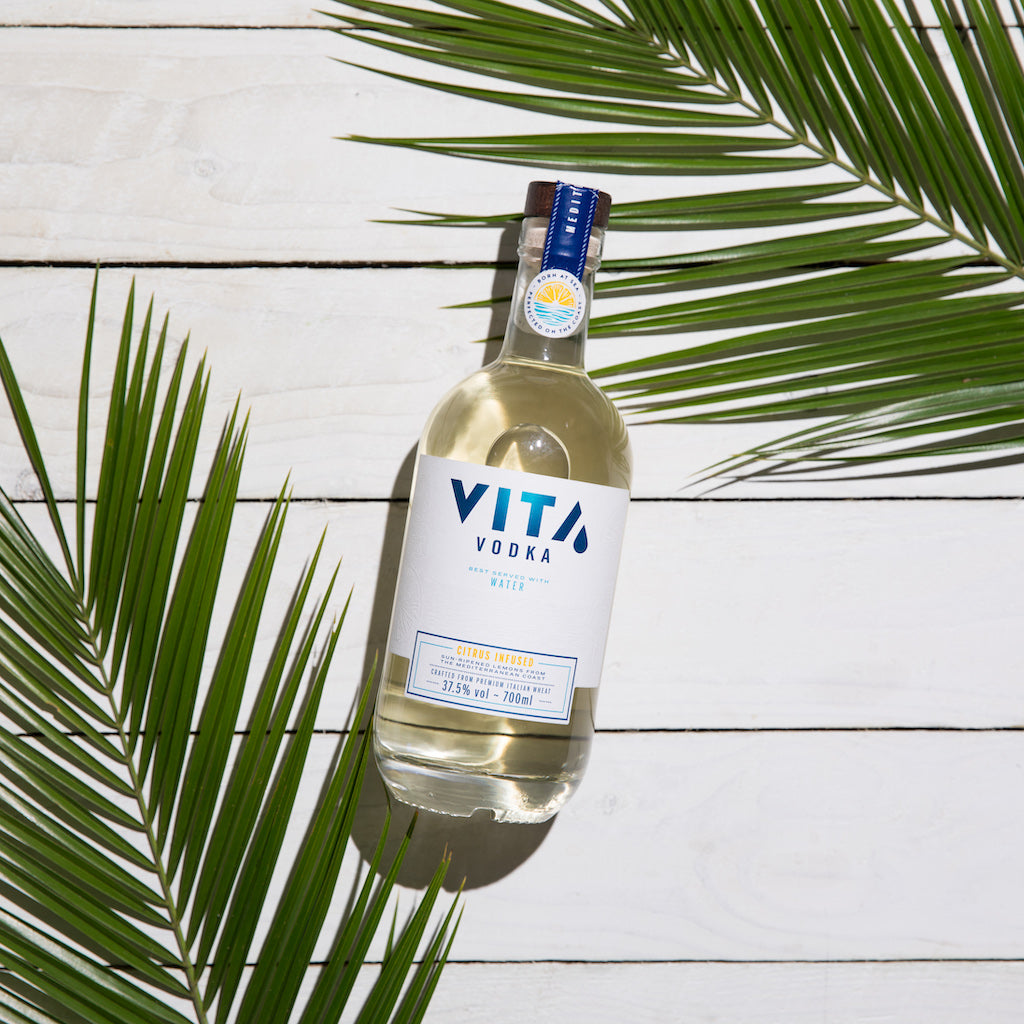 Flasche VITA Vodka auf weißem Holzhintergrund, umrahmt von grünen Palmblättern.