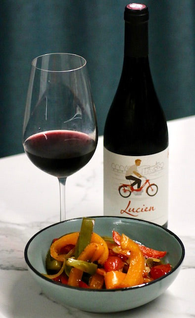 Alttext: Piperade mit Rotwein Lucien, serviert in einer blauen Schale mit einem Glas Rotwein auf weißem Marmortisch.