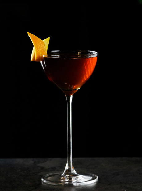 ADA Aperitif Cocktail im Coupette-Glas mit dunkler Farbe und Orangenzeste als Garnitur vor schwarzem Hintergrund.