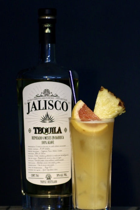 Acapulco Gold Cocktail mit Jalisco Tequila Flasche, Ananasstück und Grapefruit vor dunklem Hintergrund