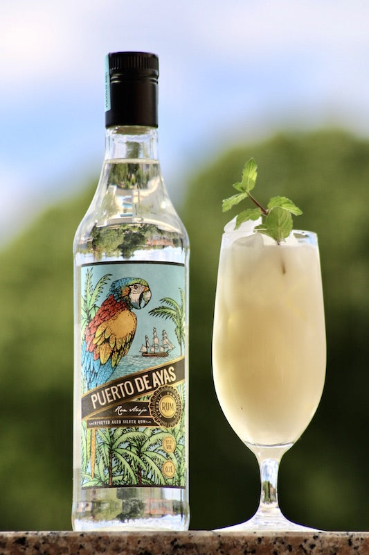 Bahia Cocktail im Glas mit Minze neben Puerto de Ayas Rumflasche mit Papagei-Etikett vor grünem, unscharfem Hintergrund