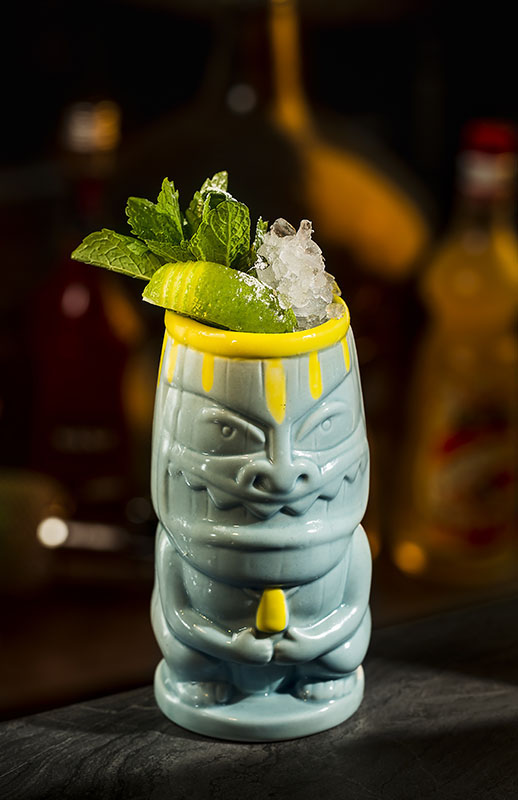 Banana Surfer Cocktail im Tiki-Becher mit Crushed Ice, Minze und Limette vor unscharfem Bar-Hintergrund