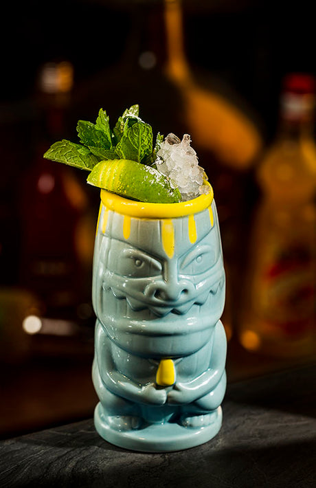 Banana Surfer Cocktail im Tiki-Becher mit Crushed Ice, Minze und Limette vor unscharfem Bar-Hintergrund