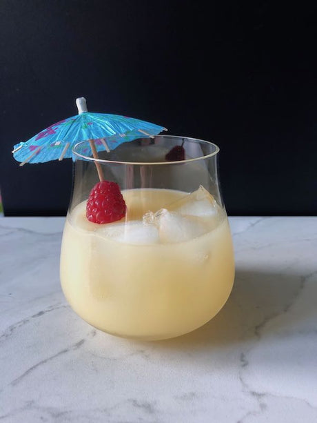 Banana de Coco Cocktail mit Eiswürfeln, Himbeere und blauem Papier-Schirmchen in einem bauchigen Glas auf Marmorfläche