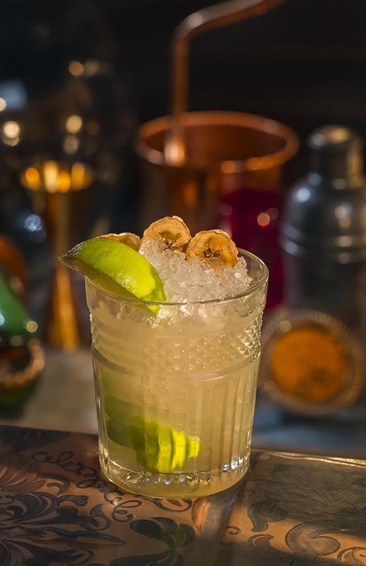 Bananen Guave Caipirinha mit Crushed Ice, Limette und Bananenchips vor Barhintergrund
