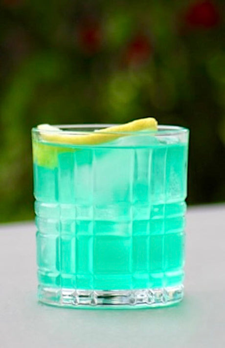 Ein erfrischender Blue Lagoon Twist Cocktail in einem Whisky-Glas, serviert mit Eiswürfeln und einer Zitronenscheibe als Garnitur. Der Cocktail hat eine leuchtend blaue Farbe und vermittelt tropisches Flair.