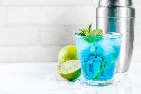 Blue'tiful: blauer Cocktail mit Eiswürfeln, Limettenzesten und Minze in einem Glas, daneben eine aufgeschnittene Limette auf Eis.