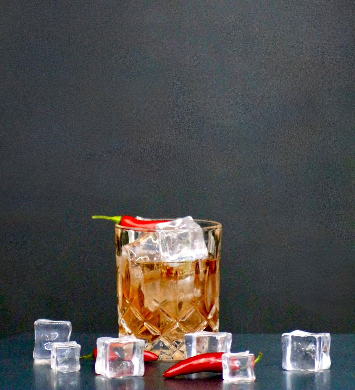 Burning Sunset Cocktail im Tumbler mit Eiswürfeln und roter Chili, umgeben von weiteren Eiswürfeln auf schwarzem Untergrund.