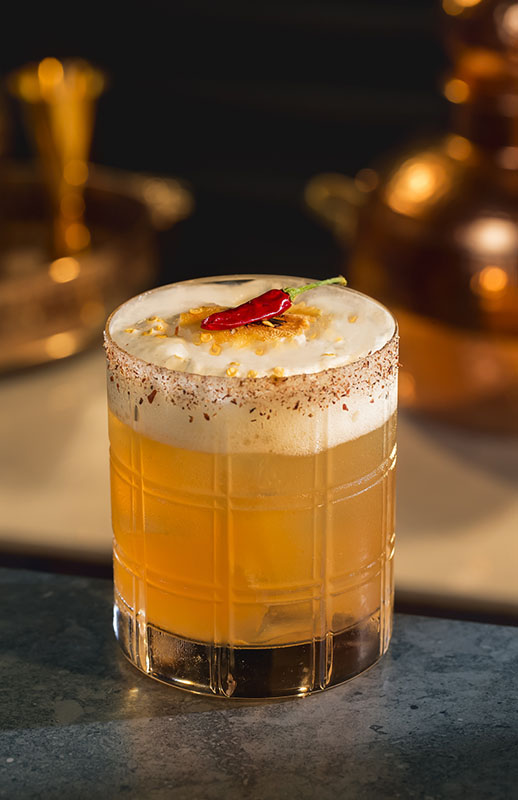 Camorra Margarita Cocktail im Rocks Glas mit Schaumschicht und Chili auf Bar vor warmem Hintergrund