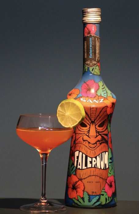 Alttext: Captain's Blood Cocktail mit Limettenscheibe, serviert vor bunter Falernum-Flasche von SANZ im Tiki-Stil